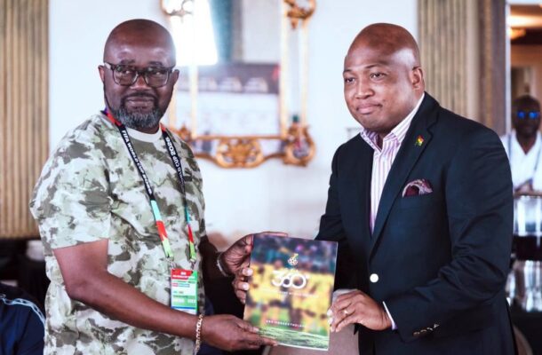 Two men in a formal setting exchange a glossy booklet or program, with the man in camouflage handing it to the man in a dark suit.