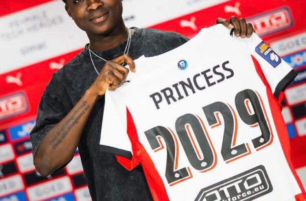 Person holding a white red-trimmed jersey that says 'PRINCESS 2029' at a sports event backdrop.