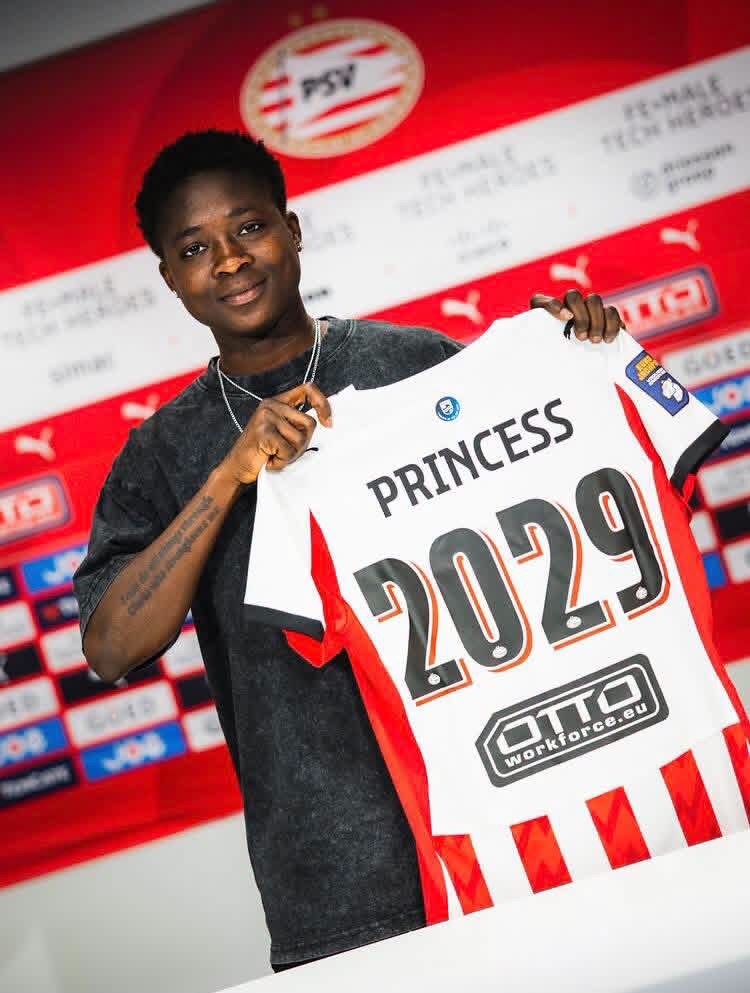 Person holding a white red-trimmed jersey that says 'PRINCESS 2029' at a sports event backdrop.