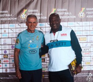 Two men posing together at a Ghana Football Association event backdrop, wearing team jerseys.