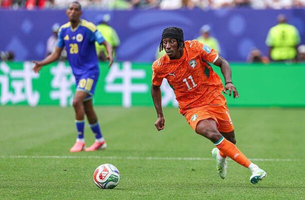 Soccer player in orange number 11 running toward the ball on a grass field, with a blue opponent close behind in the background.
