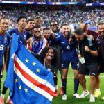A national football team posing with a flag on the field after a match, smiling and celebrating together.