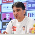 Man in a white Croatia shirt speaking into two microphones at a press conference with sponsor banners in the background.