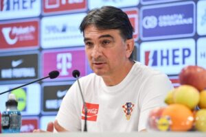 Man in a white Croatia shirt speaking into two microphones at a press conference with sponsor banners in the background.