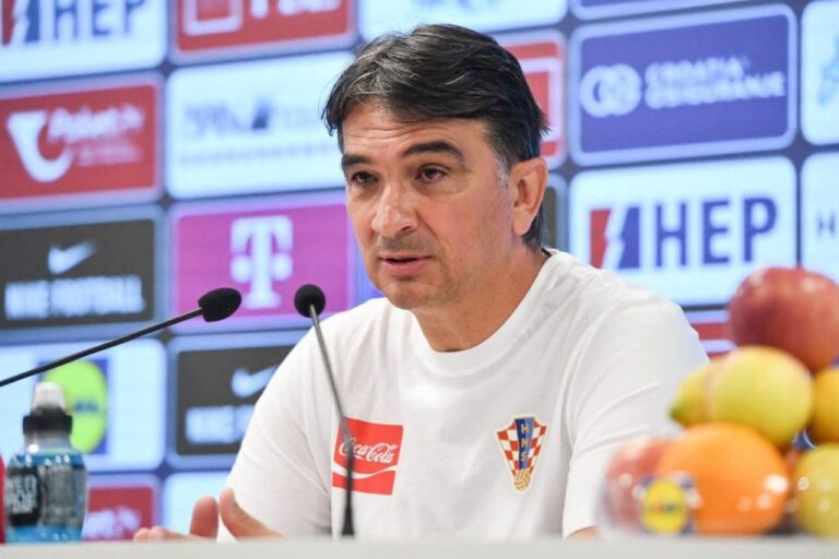 Man in a white Croatia shirt speaking into two microphones at a press conference with sponsor banners in the background.