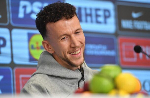 Smiling man in a gray hoodie at a press conference with sponsor logos in the background and fruit in the foreground.