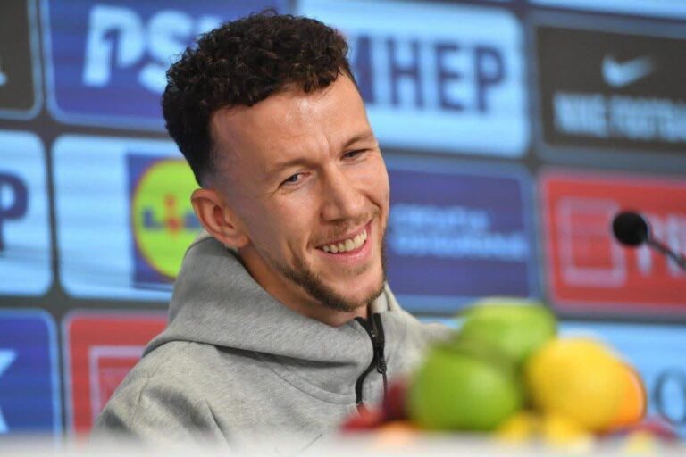 Smiling man in a gray hoodie at a press conference with sponsor logos in the background and fruit in the foreground.