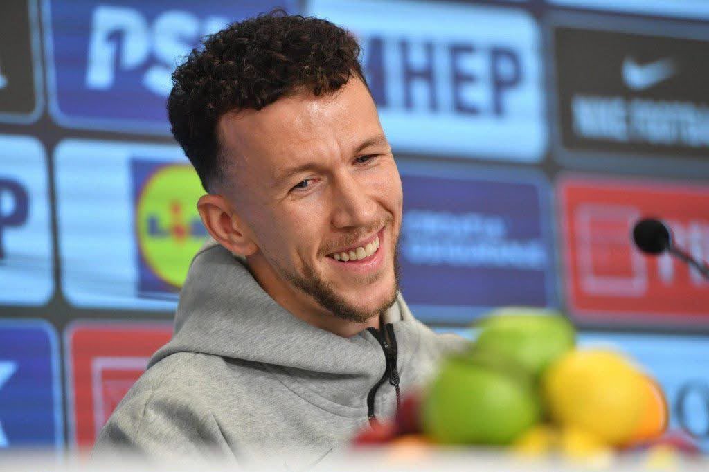 Smiling man in a gray hoodie at a press conference with sponsor logos in the background and fruit in the foreground.