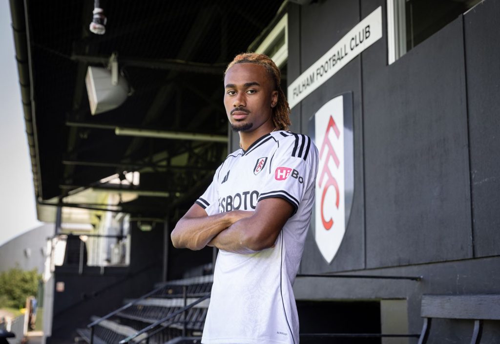 Soccer player in a white Fulham FC kit stands with arms crossed in the stadium dugout area.