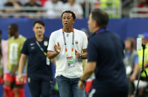Coach in a white t‑shirt with a lanyard speaks and gestures on a sports field, with players in the background.
