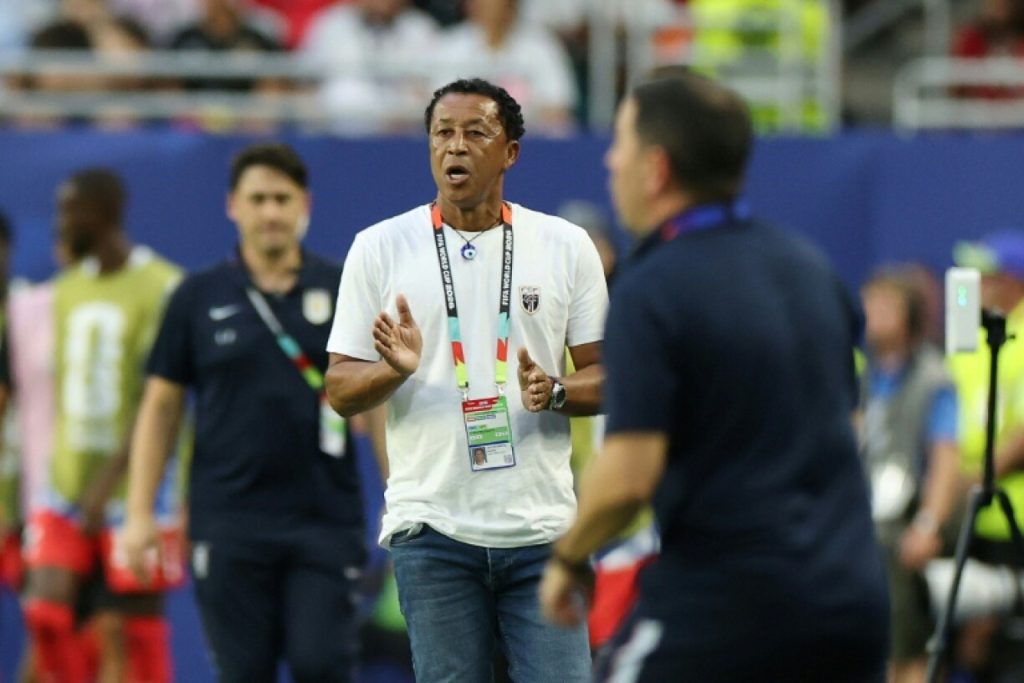 Coach in a white t‑shirt with a lanyard speaks and gestures on a sports field, with players in the background.