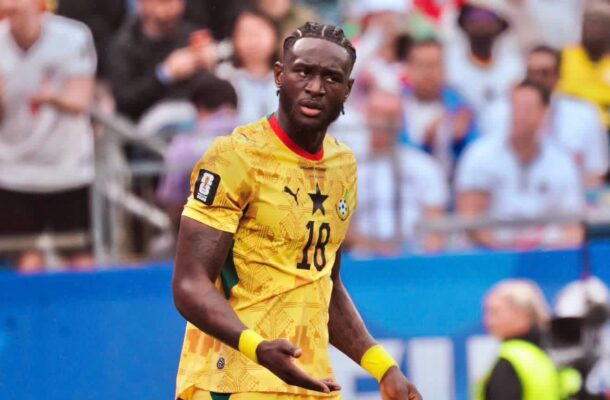 Cameroon national team footballer wearing yellow jersey with number 18, on a pitch with cheering spectators in the background.
