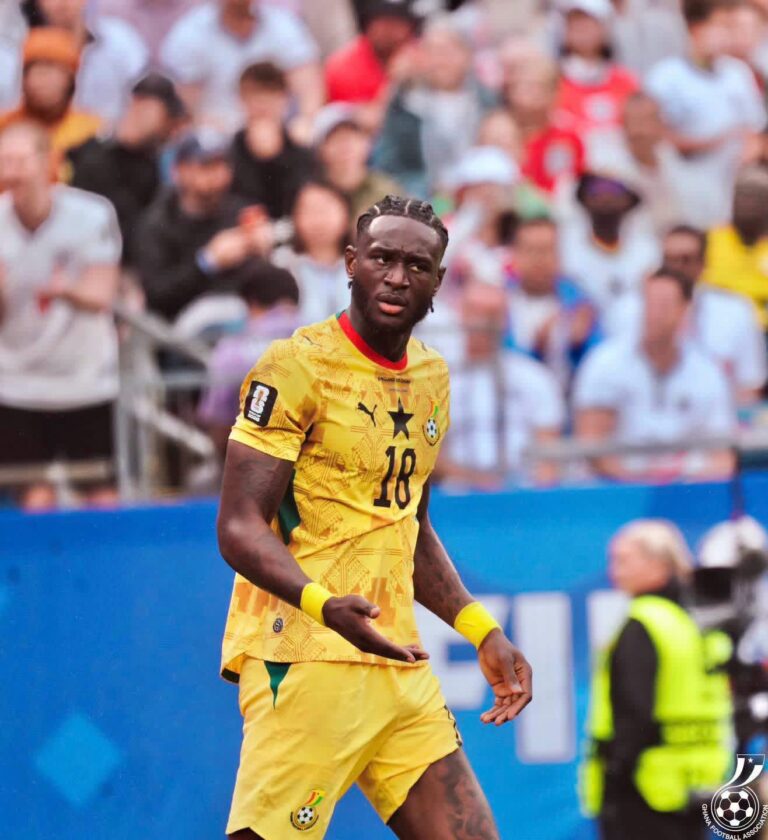 Cameroon national team footballer wearing yellow jersey with number 18, on a pitch with cheering spectators in the background.