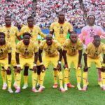 Eleven soccer players in yellow uniforms pose for a team photo on a green field with a packed stadium in the background.