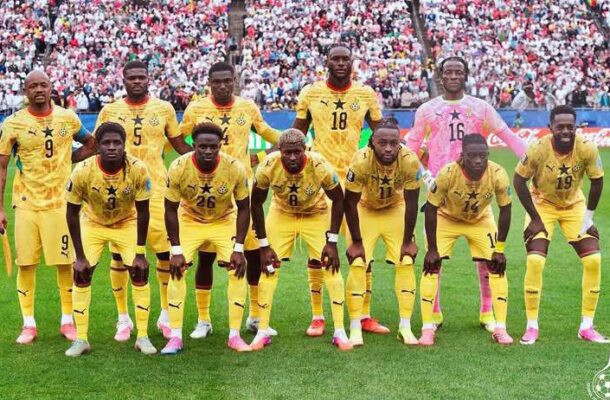 Eleven football players in yellow uniforms pose for a team photo on a grass field with a crowded stadium behind.