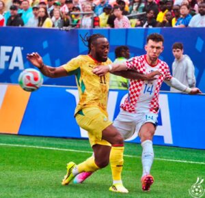 Two soccer players contest the ball during a match, one in yellow and the other in a red-and-white checker jersey, with a cheering crowd behind them.