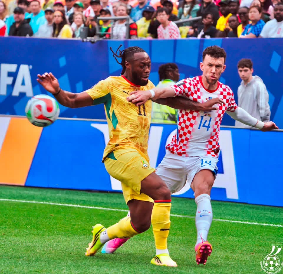 Two soccer players contest the ball during a match, one in yellow and the other in a red-and-white checker jersey, with a cheering crowd behind them.