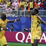 Two soccer players in yellow jerseys celebrate on the field as the crowd cheers behind them, with the word Qatar on the advertising boards.