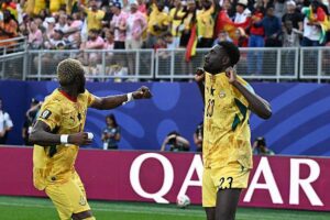 Two soccer players in yellow jerseys celebrate on the field as the crowd cheers behind them, with the word Qatar on the advertising boards.