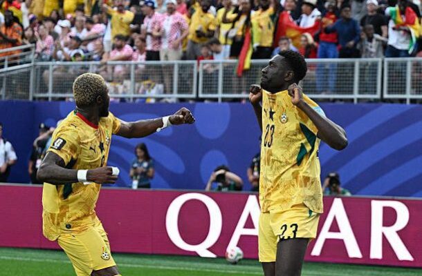 Two soccer players in yellow jerseys celebrate on the field as the crowd cheers behind them, with the word Qatar on the advertising boards.