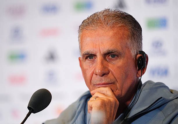 Close-up of a middle-aged man with a headset, resting his chin at a press conference microphone.”, “Man wearing a headset speaks at a press conference, thoughtful expression, microphone in front.”, “Senior man at a media event, listening intently with a microphone nearby.”, “Speaker at a press briefing, wearing an earpiece and sitting before a backdrop of logos.”