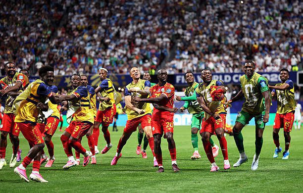 Soccer players in yellow and red jerseys celebrate on a green field as fans cheer in the stands behind them.