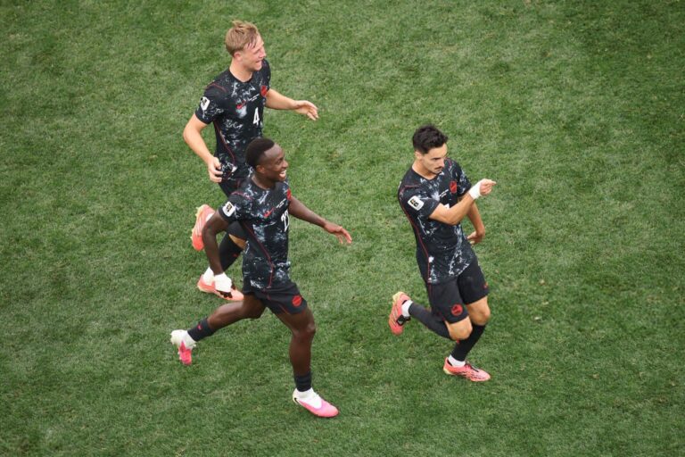 Aerial view of three soccer players in black speckled uniforms sprinting across a green grass field.