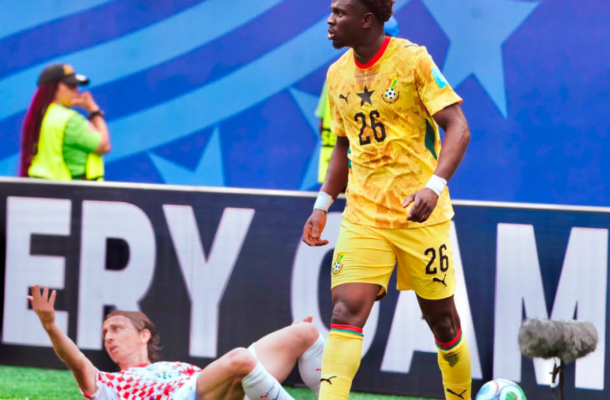 Soccer player in yellow kit (number 26) stands near a sliding opponent on the field as a ball sits nearby.