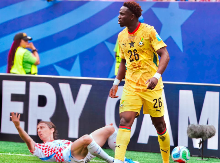 Soccer player in yellow kit (number 26) stands near a sliding opponent on the field as a ball sits nearby.