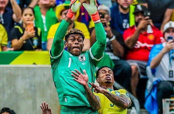 Goalkeeper in green reaches overhead to catch a soccer ball as a defender in yellow challenges him, with a cheering crowd behind.