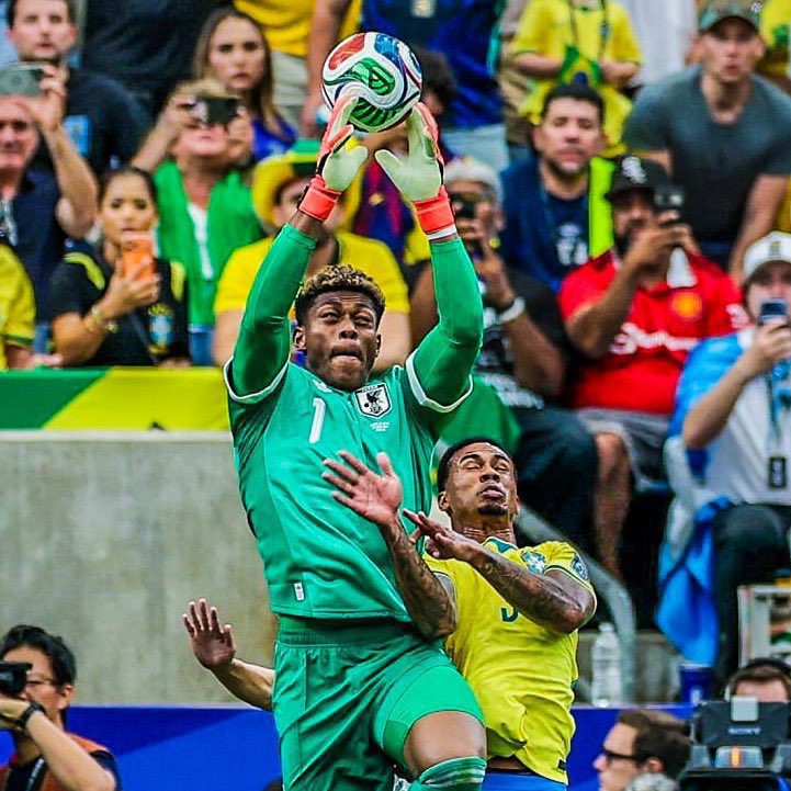 Goalkeeper in green reaches overhead to catch a soccer ball as a defender in yellow challenges him, with a cheering crowd behind.