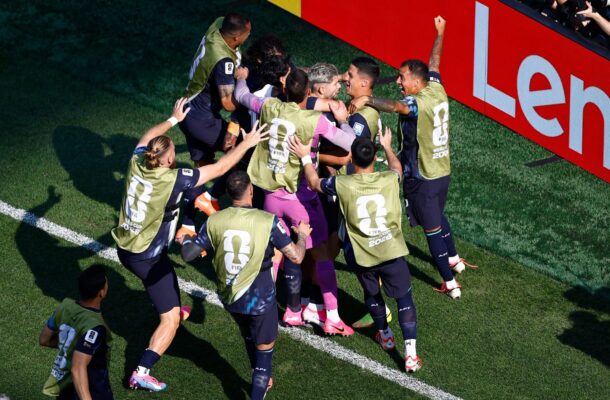 Soccer players in gold vests celebrate in a close huddle on a green field near a red advertising banner, backs showing 'FIFA World Cup 2026' logos.