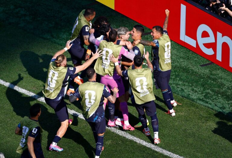 Soccer players in gold vests celebrate in a close huddle on a green field near a red advertising banner, backs showing 'FIFA World Cup 2026' logos.