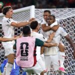 Group of soccer players in white uniforms celebrate a goal by the goal, with one player wearing a pink bib that reads FIFA World Cup 2026 in the foreground, and a net in the background.
