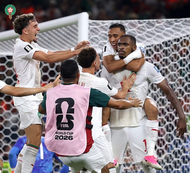 Group of soccer players in white uniforms celebrate a goal by the goal, with one player wearing a pink bib that reads FIFA World Cup 2026 in the foreground, and a net in the background.