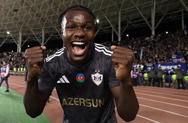 Male soccer player in a dark jersey celebrating with fists up on a stadium track, crowd cheering behind him.]? maybe too long
