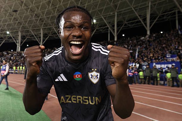Male soccer player in a dark jersey celebrating with fists up on a stadium track, crowd cheering behind him.]? maybe too long