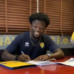 Smiling man in a navy jersey signs documents at a desk, with yellow and blue flags in the background.