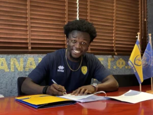 Smiling man in a navy jersey signs documents at a desk, with yellow and blue flags in the background.