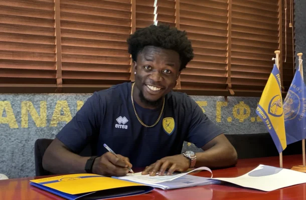 Smiling man in a navy jersey signs documents at a desk, with yellow and blue flags in the background.