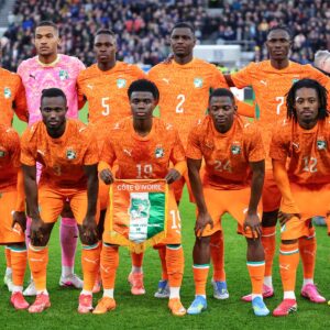 Ivory Coast national football team posing for a pre-match photo in bright orange jerseys on a green field, holding a banner reading Côte d'Ivoire.