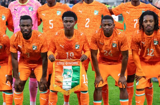 Ivory Coast national football team posing for a pre-match photo in bright orange jerseys on a green field, holding a banner reading Côte d'Ivoire.