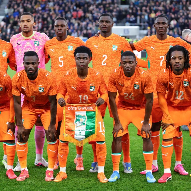 Ivory Coast national football team posing for a pre-match photo in bright orange jerseys on a green field, holding a banner reading Côte d'Ivoire.