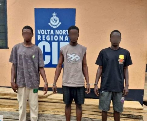 Three young men stand side by side with their wrists restrained in front of a pale orange wall and a blue sign reading 'Volta North Regional'—likely at a police facility.