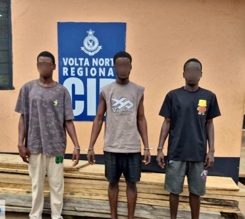 Three young men stand side by side with their wrists restrained in front of a pale orange wall and a blue sign reading 'Volta North Regional'—likely at a police facility.