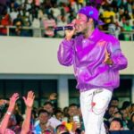 Male performer in a purple jacket and cap singing into a microphone on a crowded stage. Audience raises hands in excitement below him.