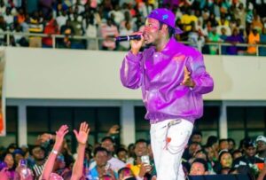 Male performer in a purple jacket and cap singing into a microphone on a crowded stage. Audience raises hands in excitement below him.