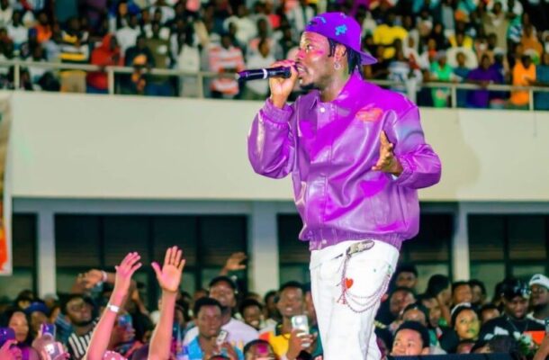 IMG_7003-779x1024 | Ghanaguardian.com Male performer in a purple jacket and cap singing into a microphone on a crowded stage. Audience raises hands in excitement below him.