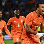 Three Senegal national team players in orange kits celebrate a goal on the soccer field, foreground player shouting with arms outstretched.