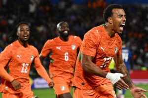 Three Senegal national team players in orange kits celebrate a goal on the soccer field, foreground player shouting with arms outstretched.